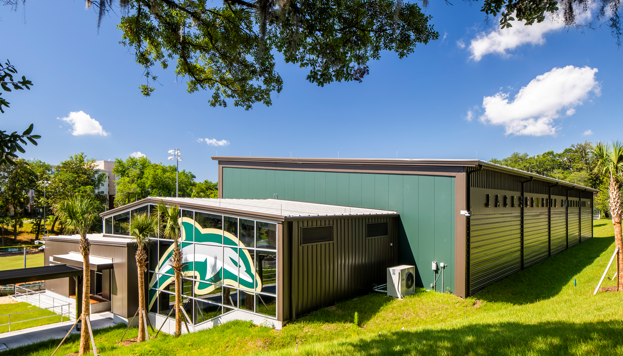 Green metal exterior of the Jacksonville University Basketball Performance Center featuring a large dolphin mascot graphic and covered walkway.