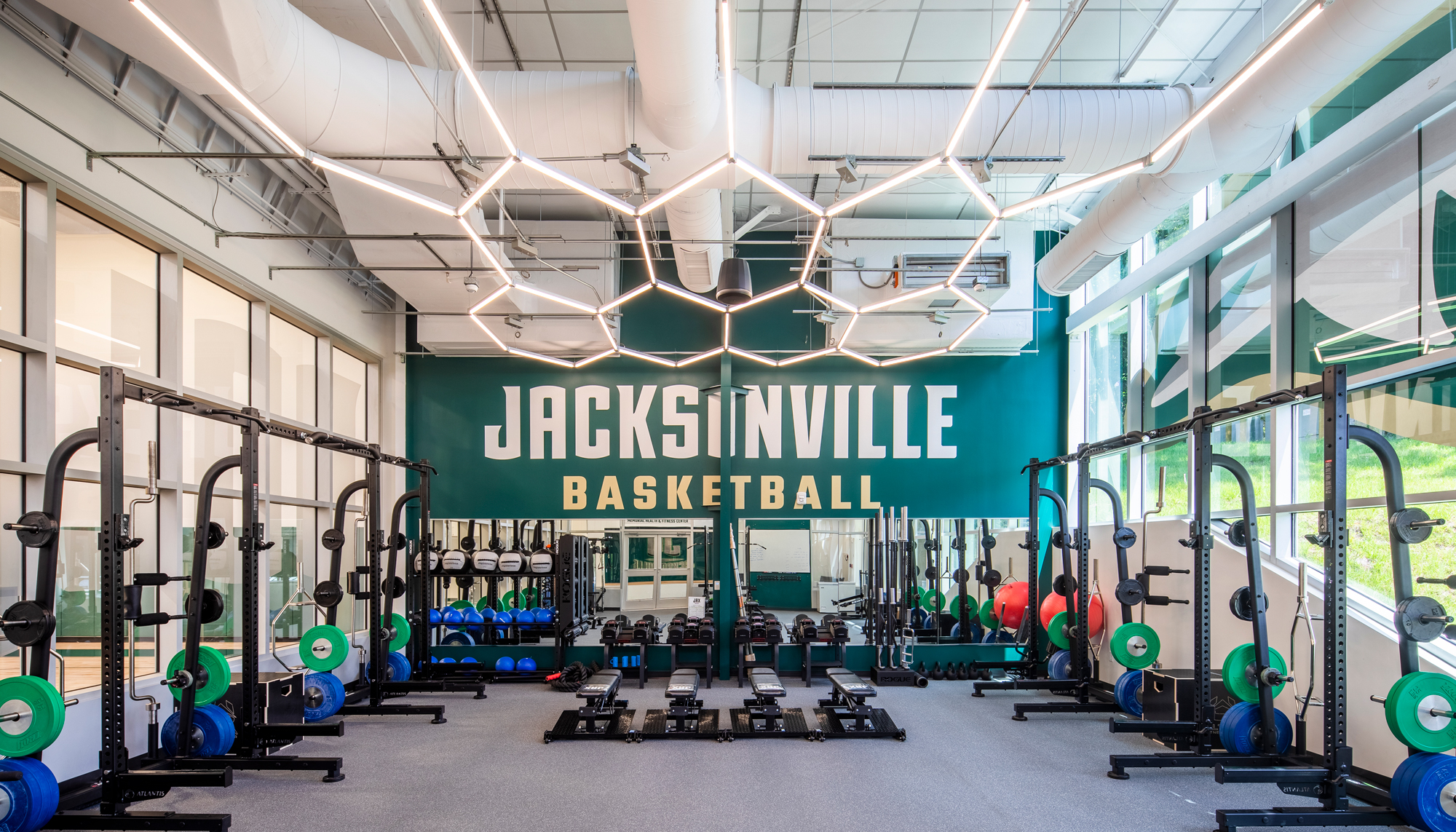 Interior strength training area at the Jacksonville University Basketball Performance Center featuring weightlifting equipment, geometric ceiling lights and a large teal wall with Jacksonville Basketball lettering.