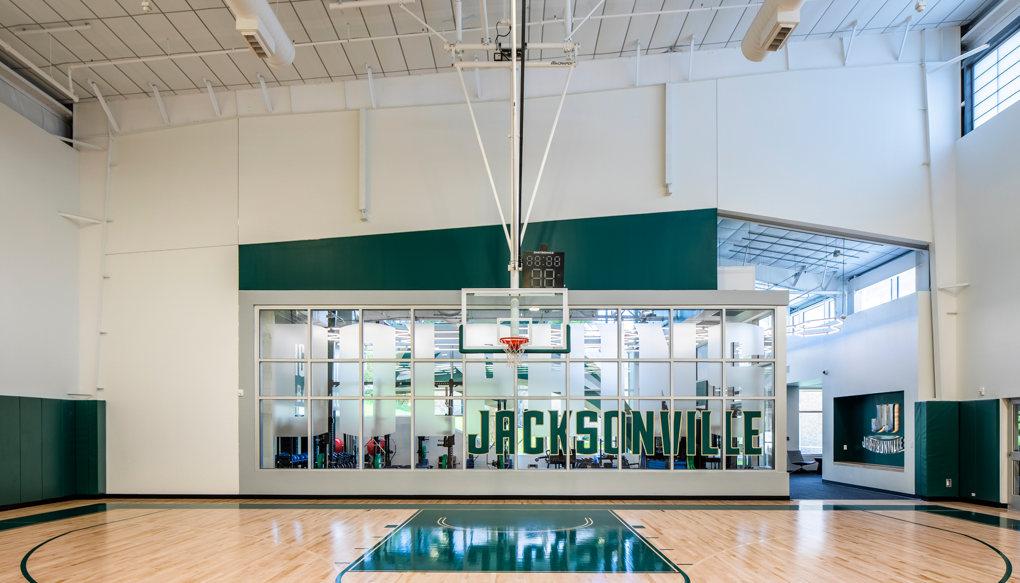 Indoor basketball court at the Jacksonville University Basketball Performance Center featuring an angular recessed wall design, glass training rooms and green Jacksonville lettering.