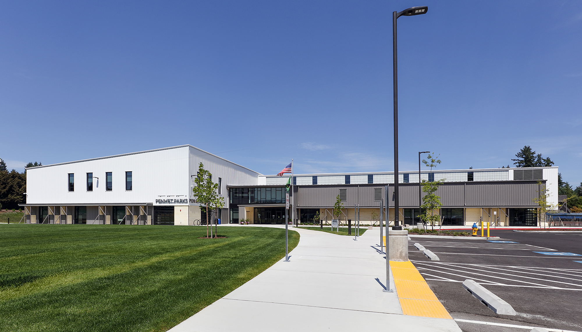 Modern white and gray community building with a green lawn, wide sidewalk, parking lot and black light poles under a clear blue sky.
