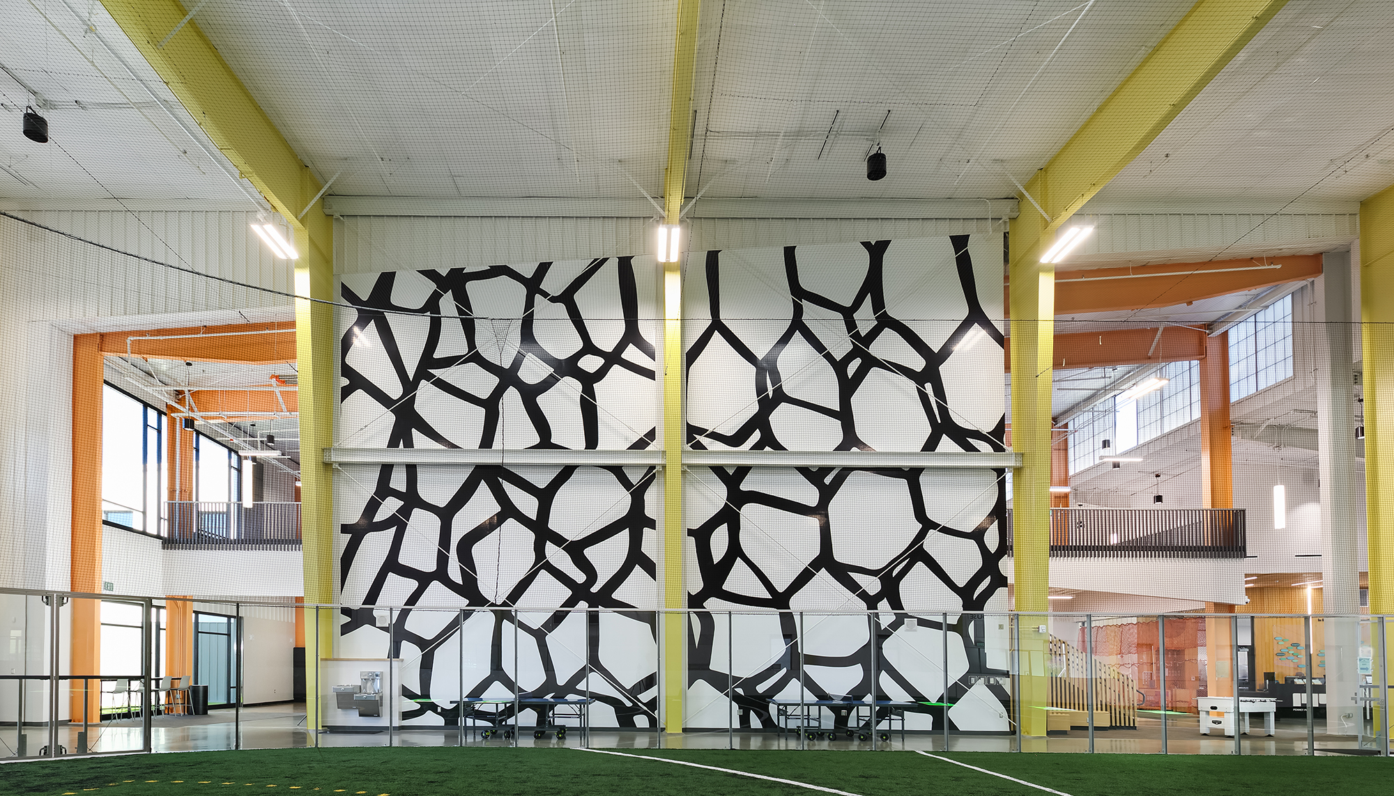 Bright recreation facility interior with green turf, yellow steel columns, orange accents and a large black-and-white abstract wall graphic.