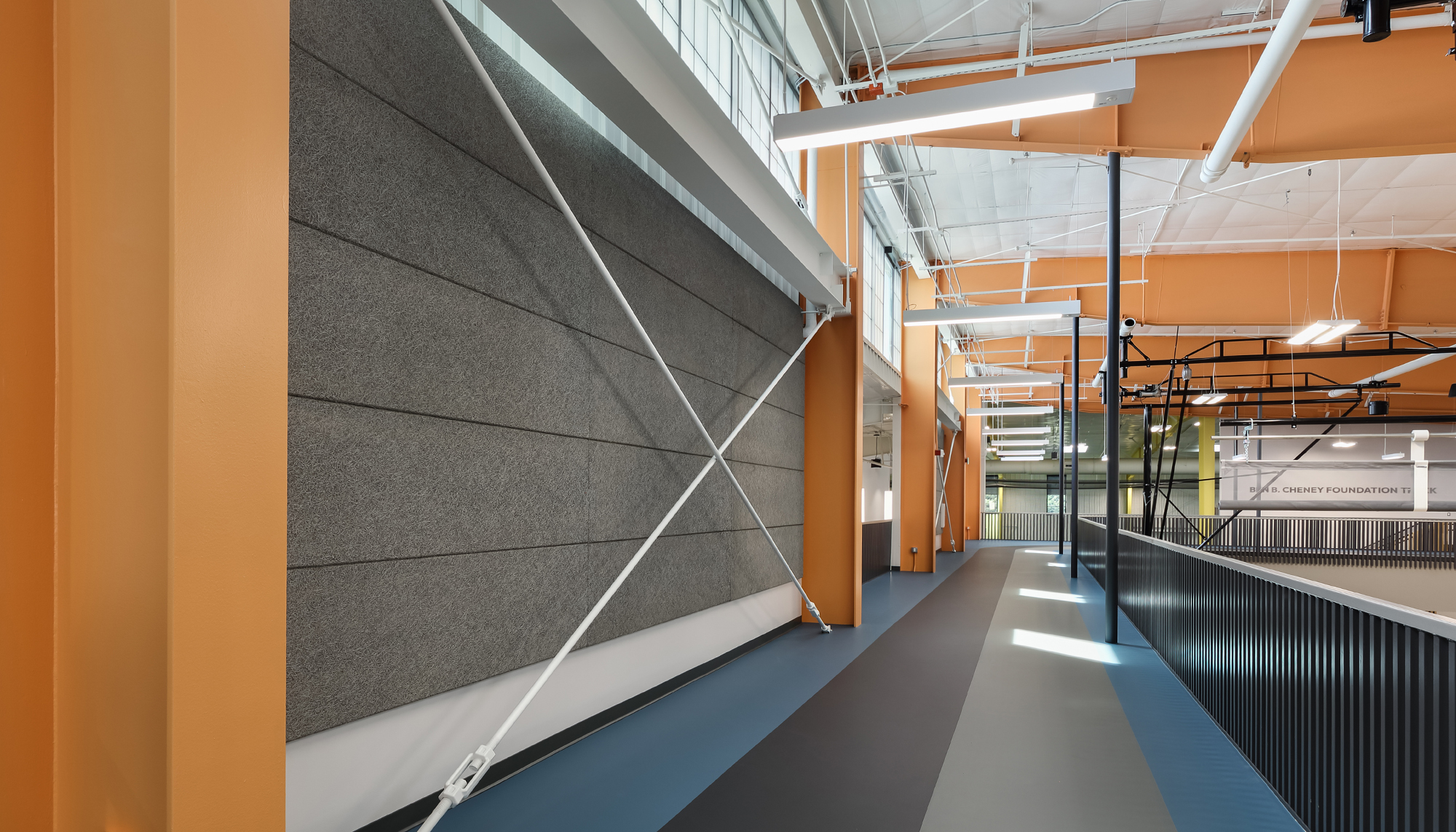 Modern interior walkway with orange steel columns, gray acoustic wall panels, blue and gray flooring and black railings beneath an exposed white ceiling.