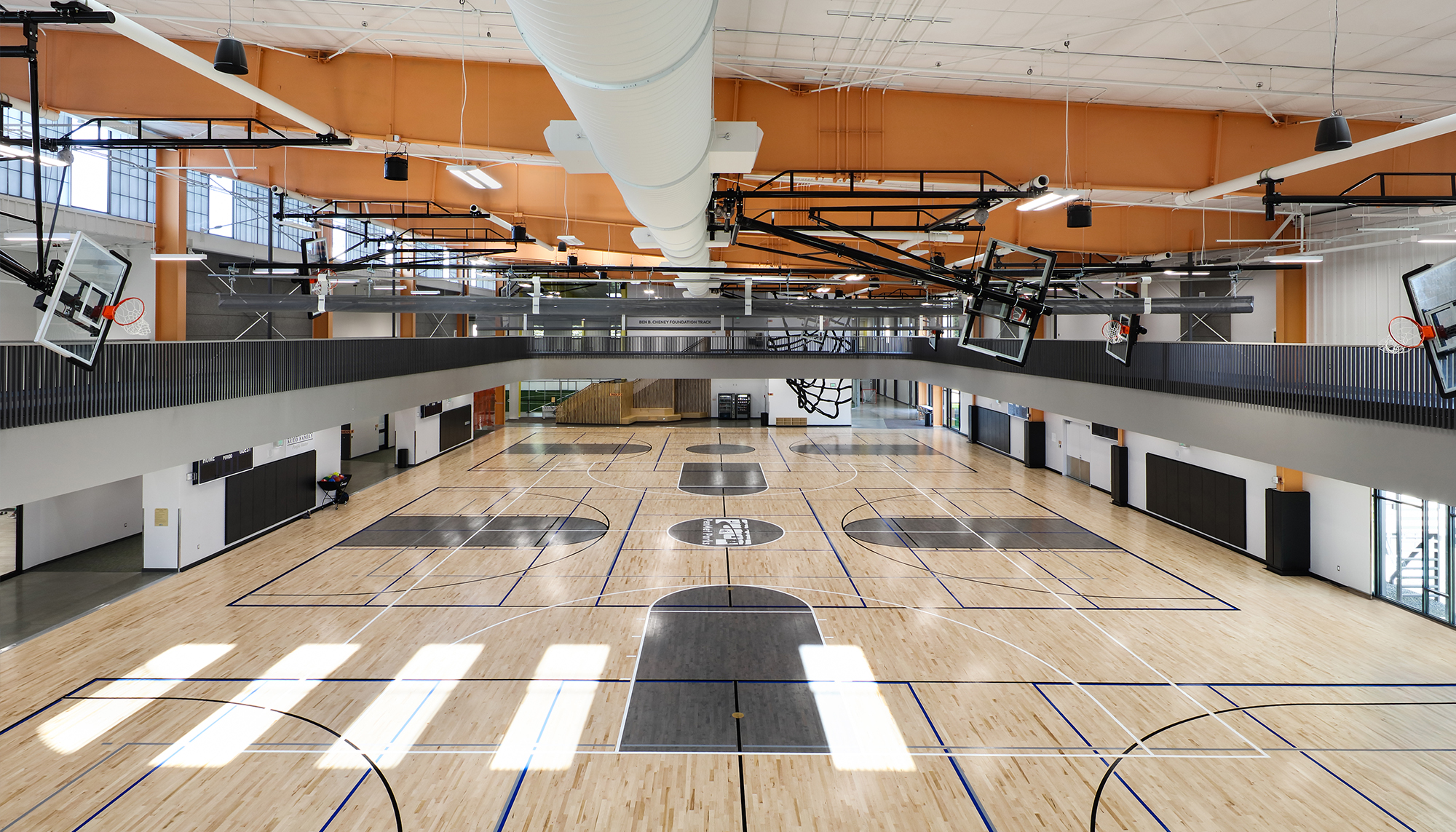 Indoor basketball facility with light wood courts, black markings, orange steel beams, white ductwork and multiple suspended basketball hoops.