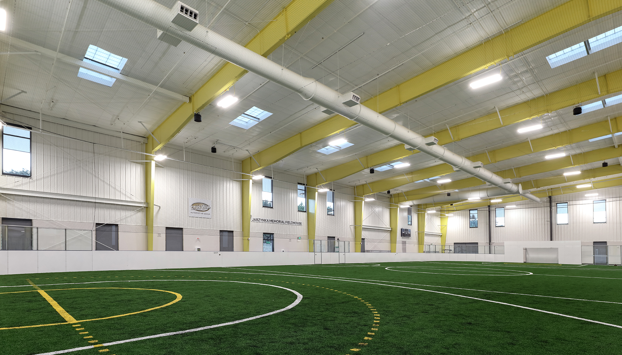 Indoor sports facility with green artificial turf, white walls, yellow steel beams, white ductwork and bright overhead lighting.