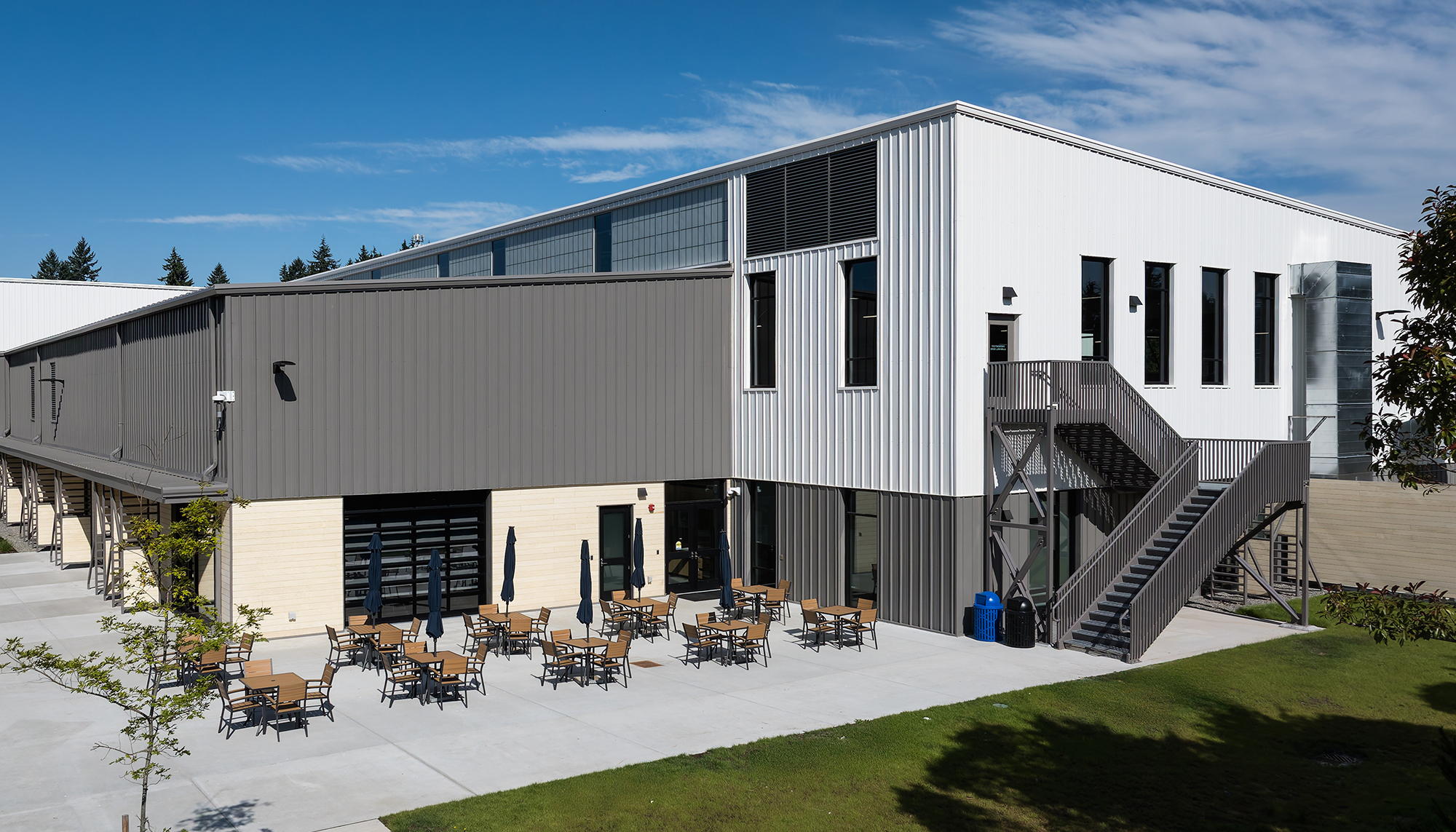 Modern gray and white metal building with an exterior staircase, black-framed windows and an outdoor patio with tables and chairs.