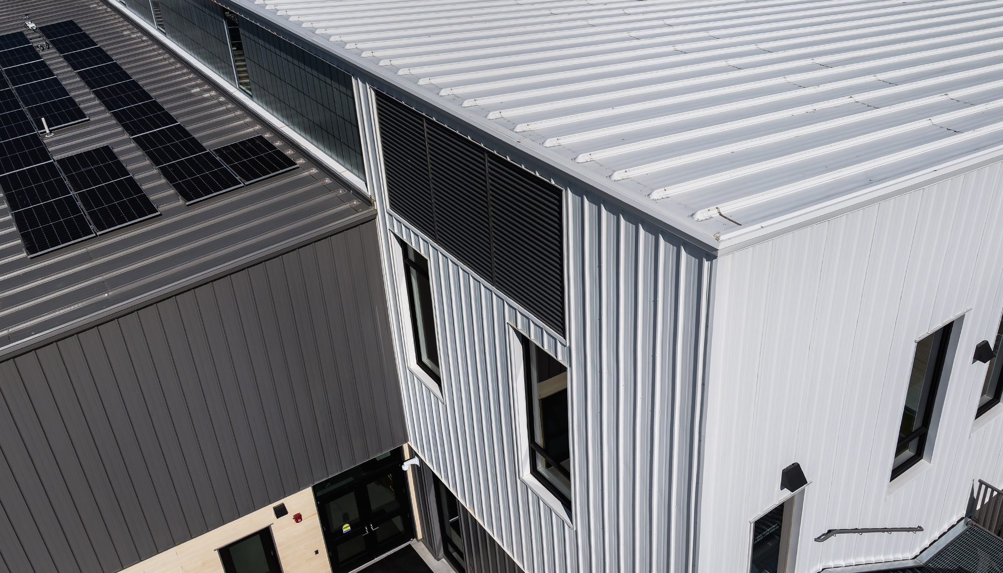 Aerial view of a gray and white metal building with ribbed roof and wall panels, black-framed windows and rooftop solar panels.