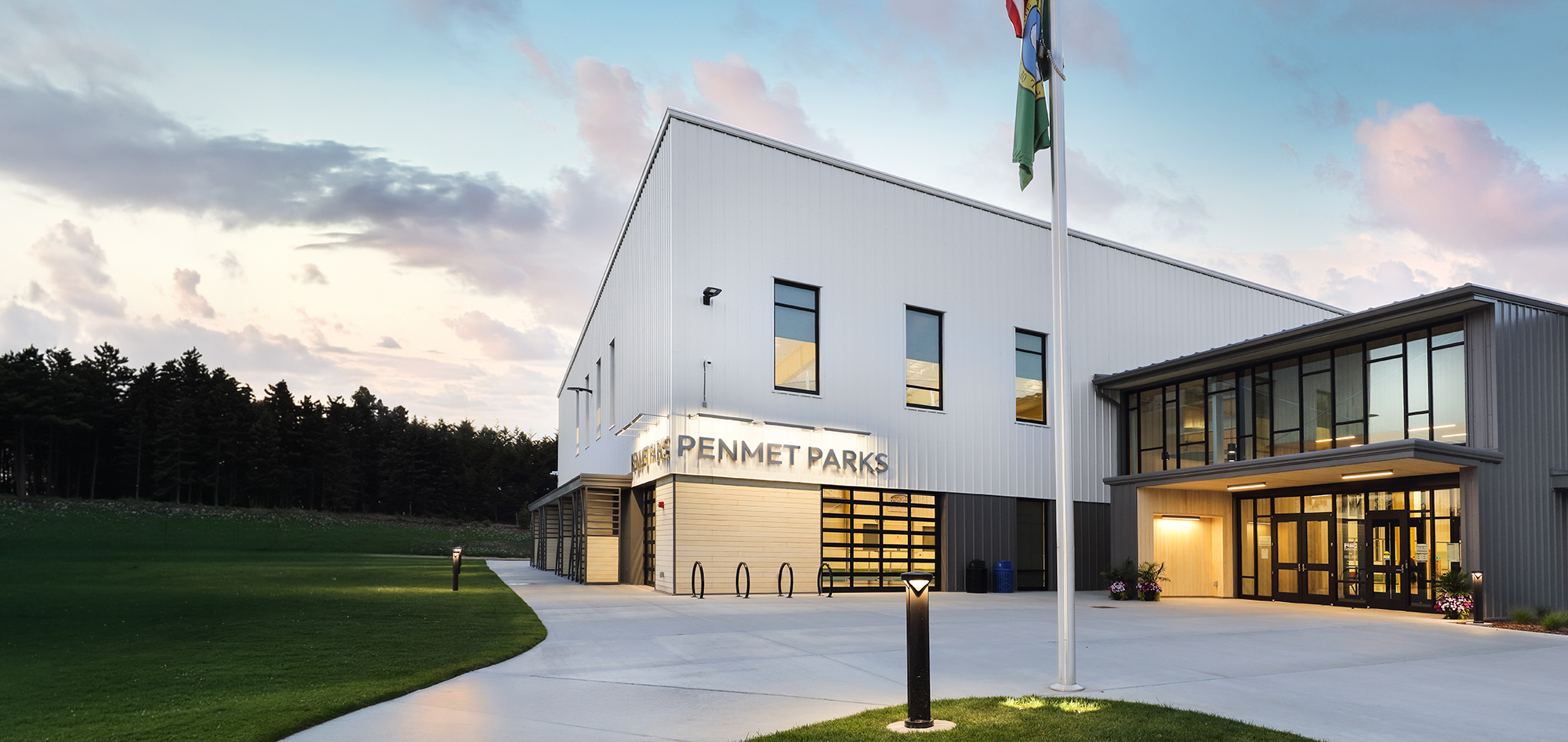 ALT text: Modern white and gray Kempenfelt Parks building with large windows, warm interior lighting, green lawn and a flagpole at dusk.