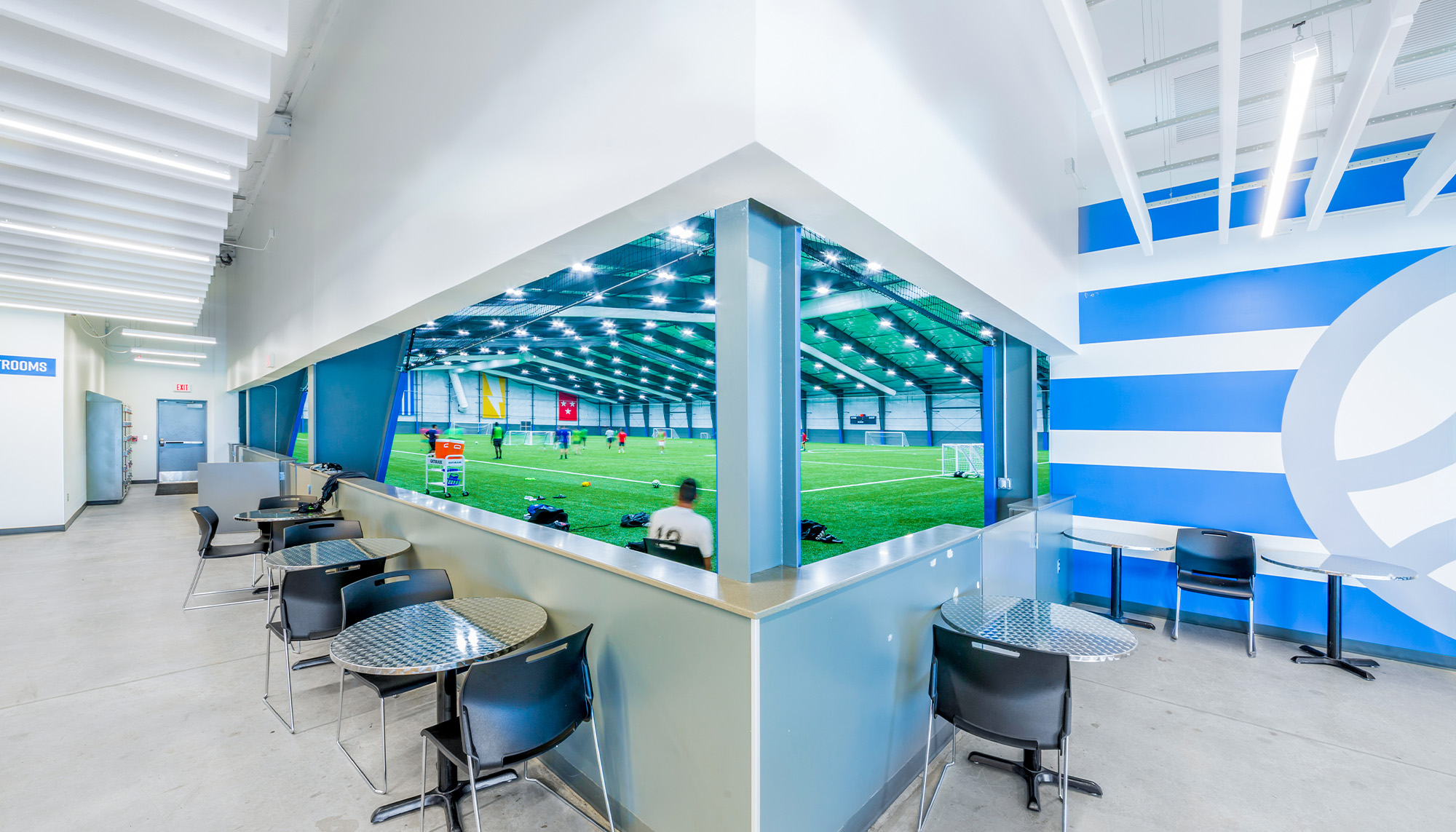 Indoor sports complex lounge featuring spectator viewing area, clear span steel framing and synthetic turf athletic field.