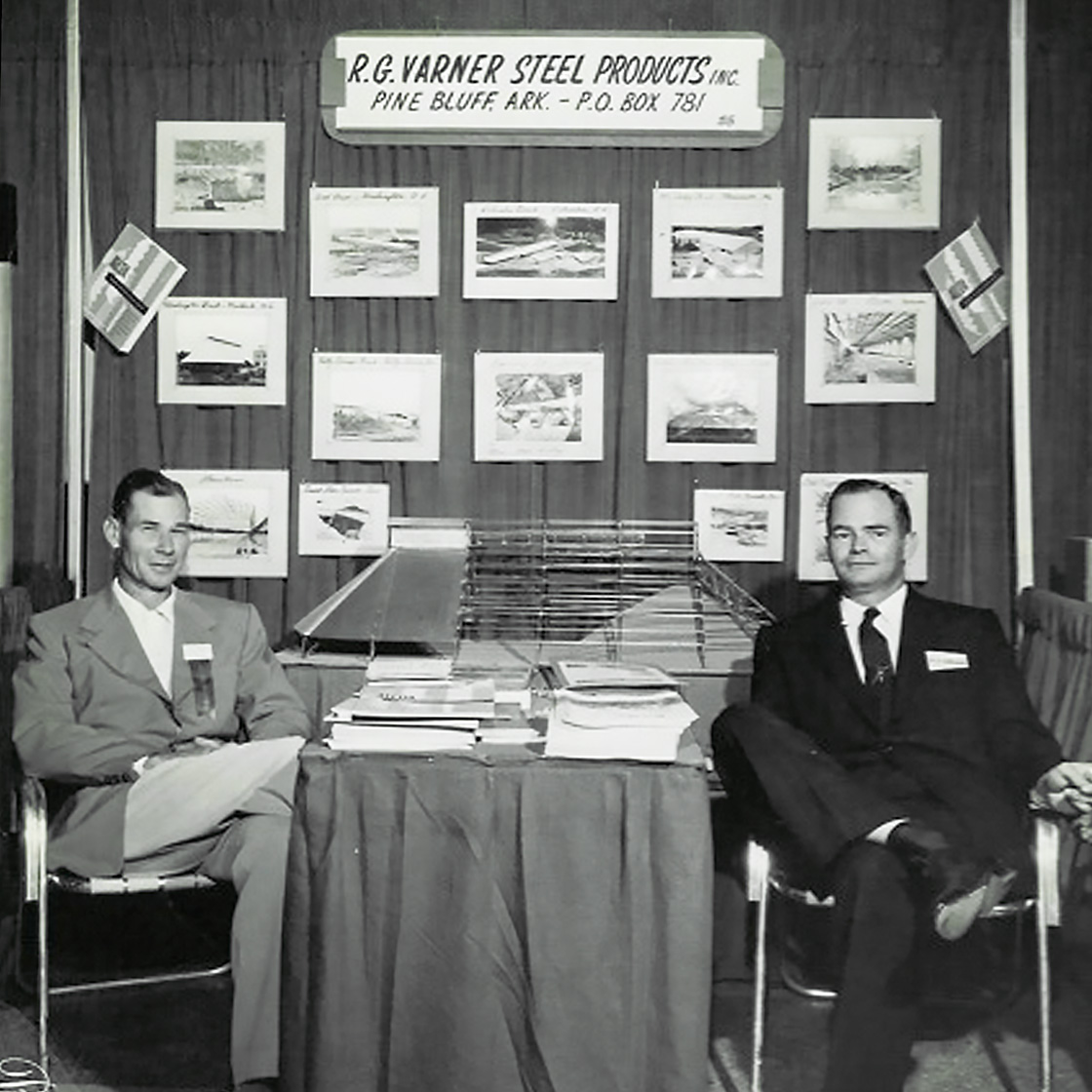 Historic black-and-white photograph of early steel building pioneers seated beside a scale building model and project display, representing the origins of steel building innovation in Arkansas.