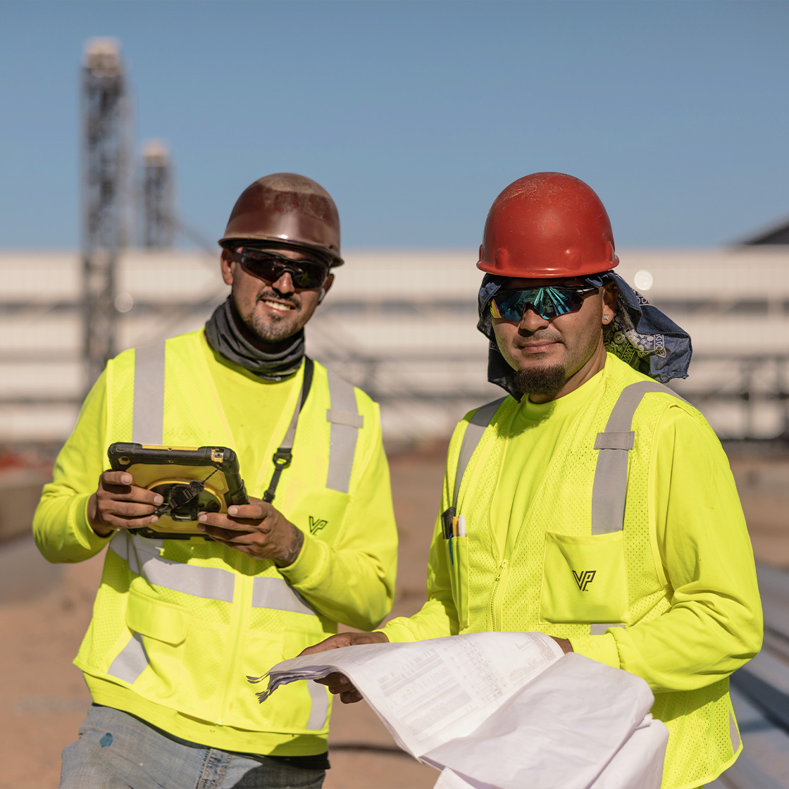 Two construction professionals in high-visibility safety gear reviewing project plans and digital site data on an active industrial job site.