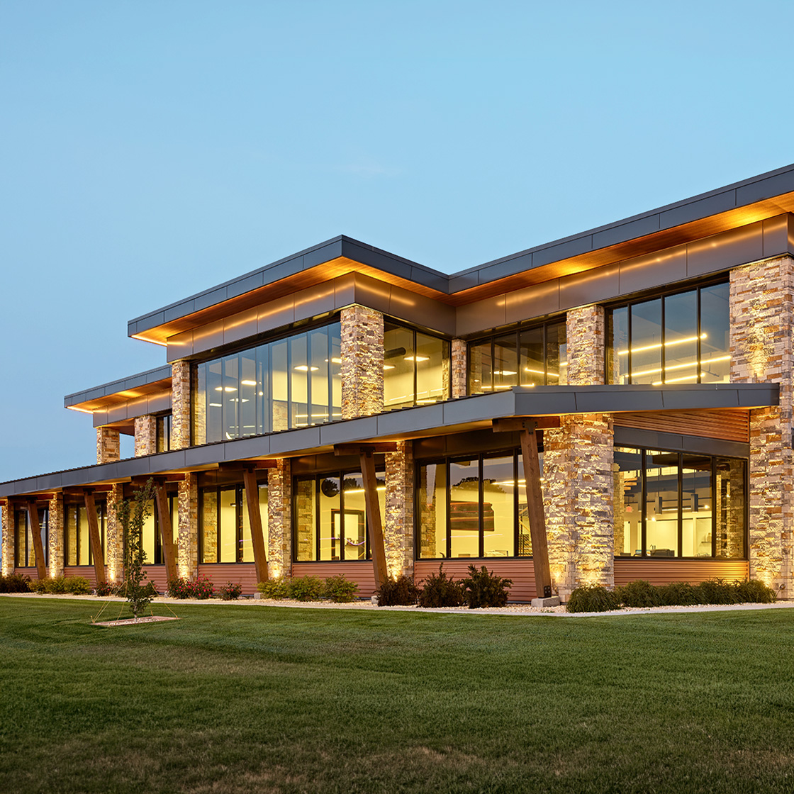 Contemporary two-story office building with illuminated glass walls, stacked stone columns and extended roof overhangs at dusk.