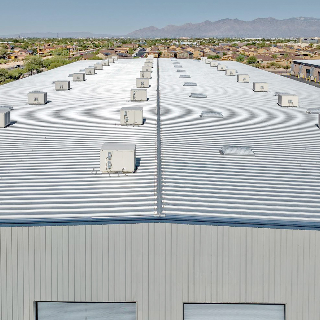 Expansive white standing seam roof system with evenly spaced rooftop units atop a large industrial facility in a suburban landscape.