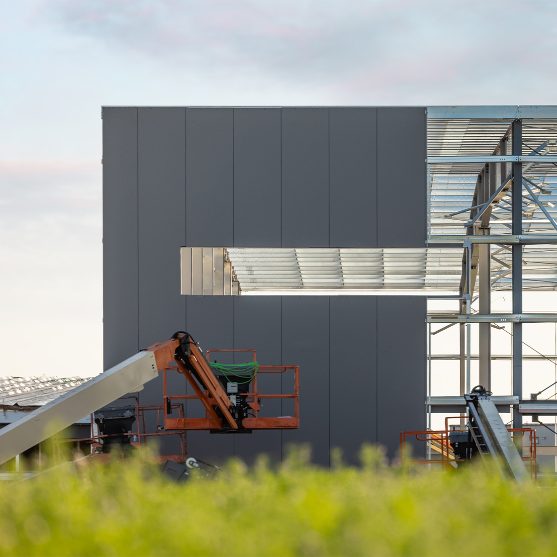 Steel building under construction with dark gray metal panel installation, exposed structural framing and elevated work platforms on site.